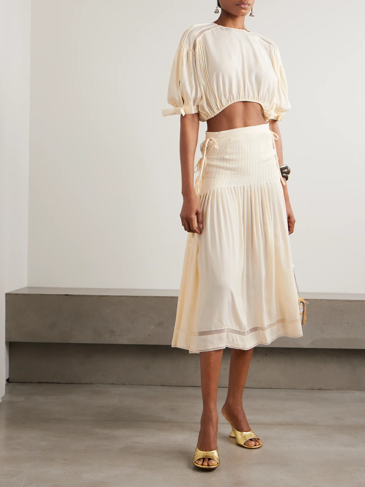 Image of a model wearing Tops. Product name - Cropped Bow-detailed Lace-trimmed Pleated Crepe Top in Ecru colour by brand - RABANNE
